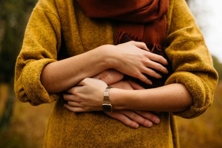 Geborgenheit fühlen: Eine Frau in einem senfgelben Pulli und rotem Schal wird von hinten von einer lieben Person umarmt