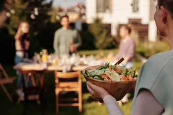 Mehr Ruhe im Alltag: Nachbarschaft pflegen. Zu sehen ist ein Gartenparty, eine Frau bringt eine Schüssel mit Salat zu einem gedeckten Tisch im Garten.