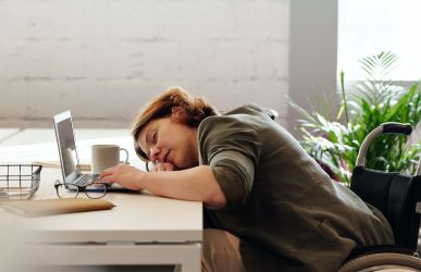 Ursachen von Erschöpfung und Burnout: Frau im Rollstuhl an einem Schreibtisch, legt erschöpft den Kopf auf die Tischplatte vor ihrem Laptop