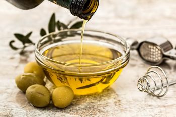 Lebensmittel gegen Entzündungen: Olivenöl. Zu sehen ist eine Schale, in die Olivenöl gegossen wird. Zur Deko liegen Oliven und Küchenutensilien um die Schale