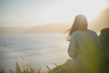 Immunsystem natürlich mit Vitamin D stärken: eine Frau sitze auf einem Hang und schaut auf das Meer. Die Haare wehen im Wind, die Sonne scheint am Horizont