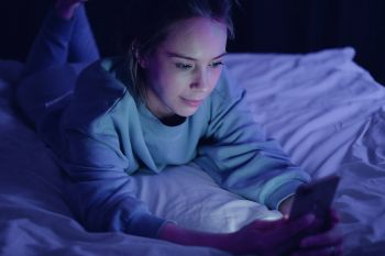 Blaues Licht: Schlaf. Zu sehen ist eine junge Frau, die auf ihrem Bett liegt und auf ihr Handy schaut.